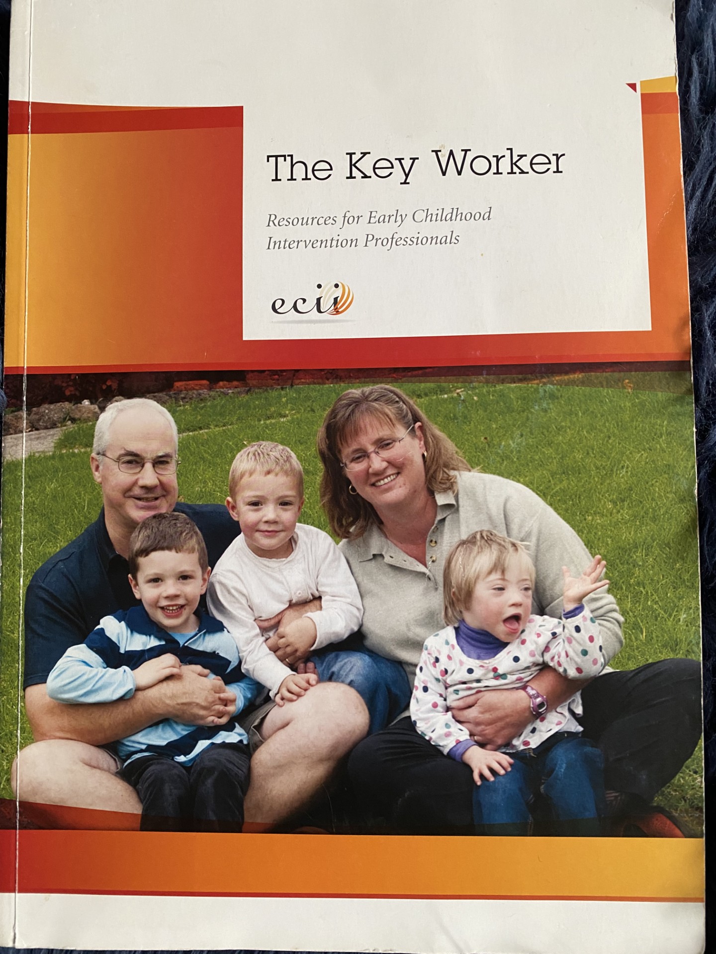 Words - The Key Worker, Resources for Early Childhood Intervention Professionals. Letters - ecii. Photograph of a family of five, sitting cross-legged on grass. Two young boys sitting on their father's lap and a small girl sitting on her mother's lap.