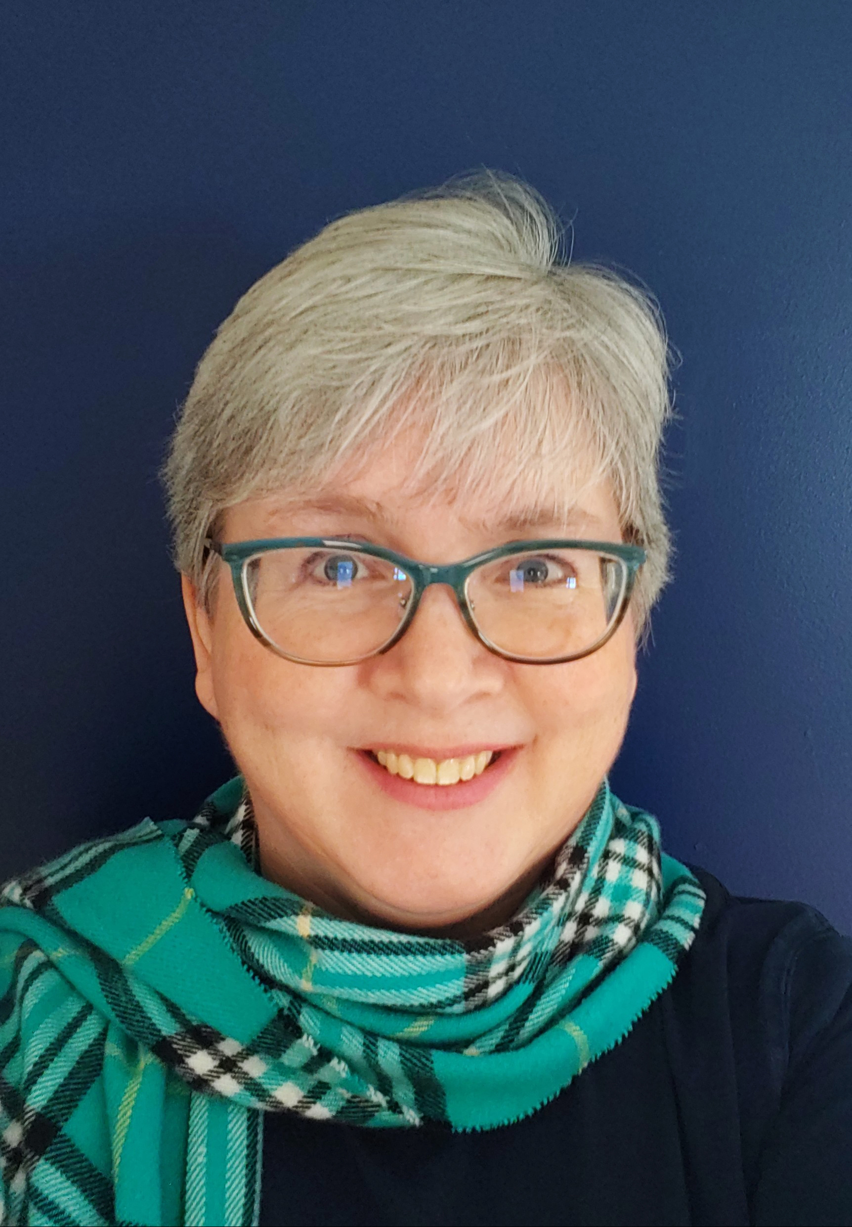 Smiling woman with short gray hair, a turquoise plaid scarf and turquoise framed glasses.