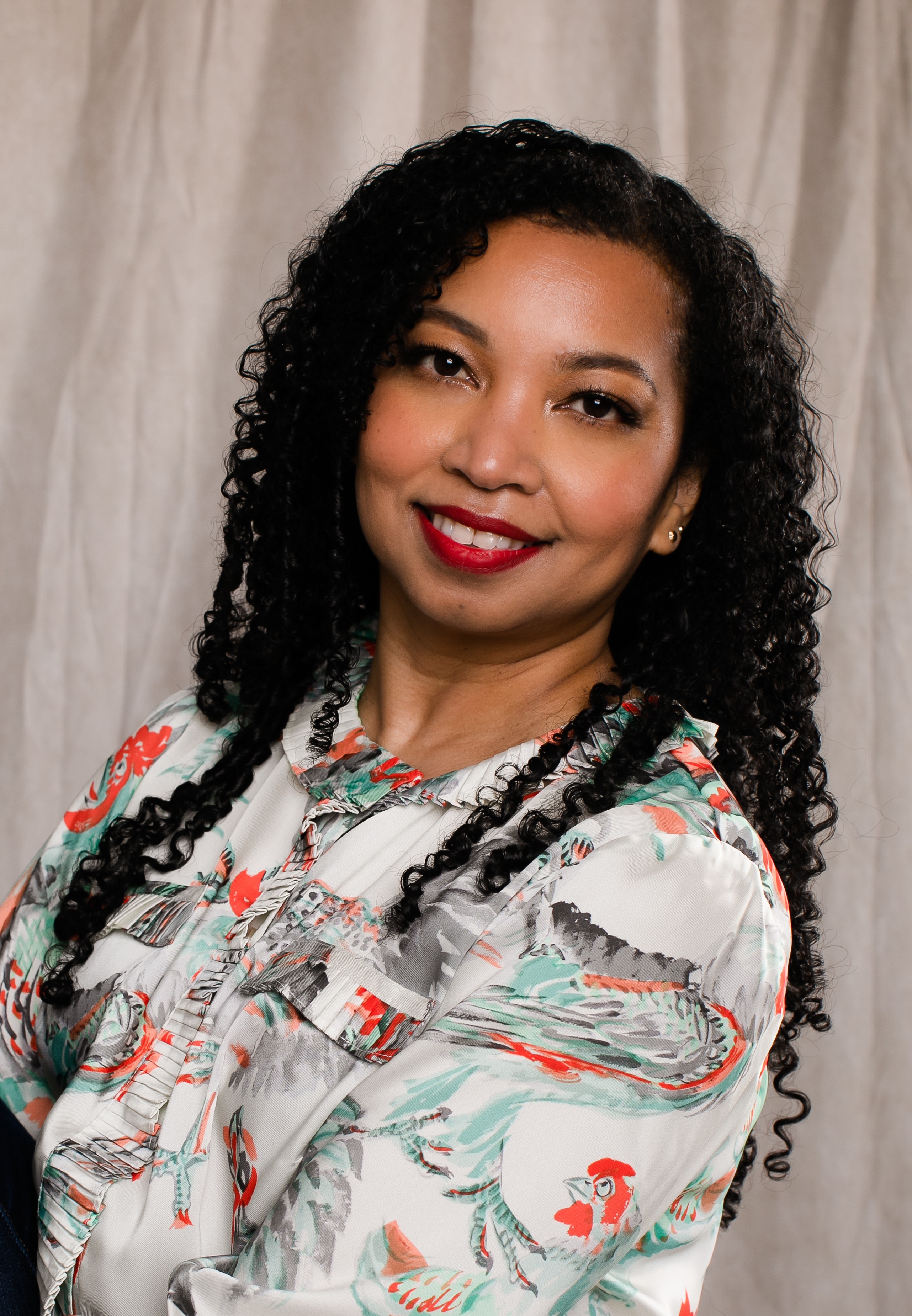Photo of Black woman with long back curly hair, wearing a print shirt in white, blue and orange.