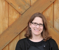 Kathy MacMillan smiles into the camera. She is a White woman with light brown, shoulder-length hair, brown eyes, and glasses. She wears a black top with a black cardigan over it. She is sitting against a wooden door.