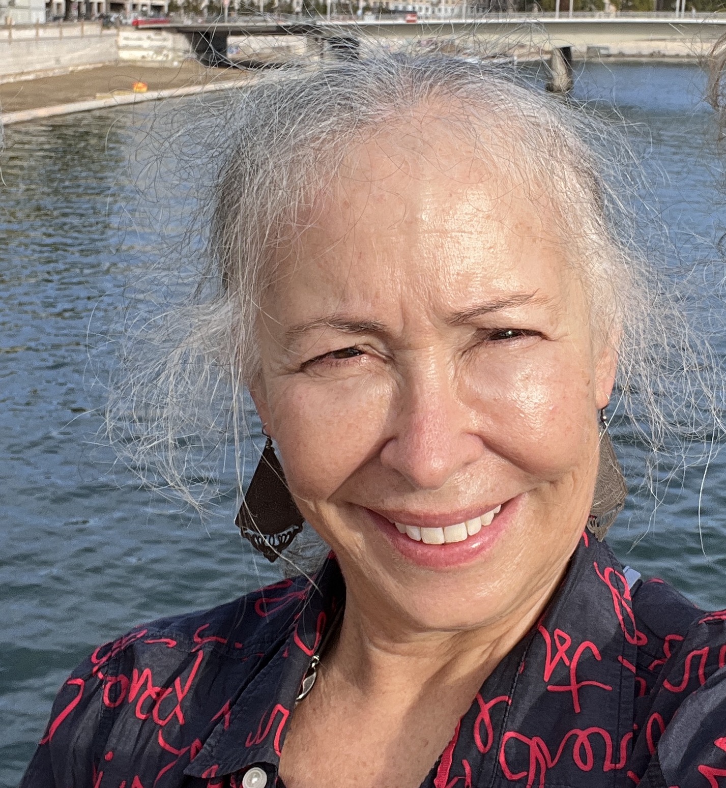 selfie of Ms. Leslie taken in Annecy, France as she squints facing the sun with backdrop of bridge over river