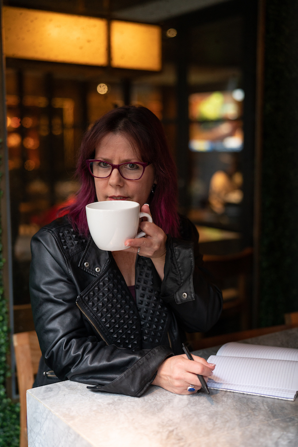 A woman wearing glasses seated at a table with a coffee mug and notebook.