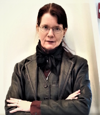 Middle-aged person with glasses wearing black leather jacket and waistcoat over burgundy silk shirt, red hair in a chignon; standing indoors against a white wall