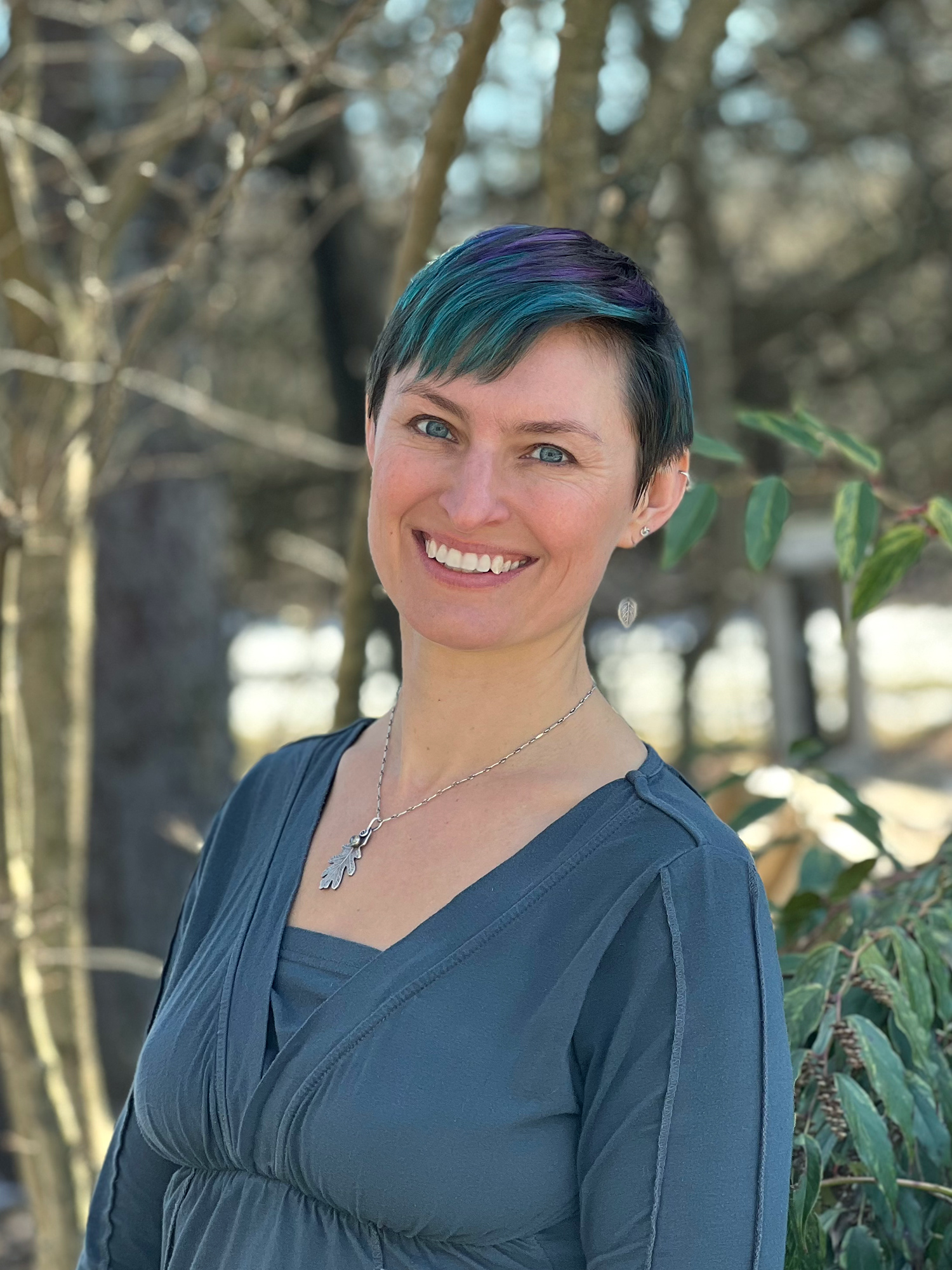Author and illustrator Victoria K. Chapman stands outside by a stand of trees and smiles. She has a short pixie style haircut that has turquoise and purple in it, blue eyes, and light tan skin. She is wearing a grey long sleeve shirt.