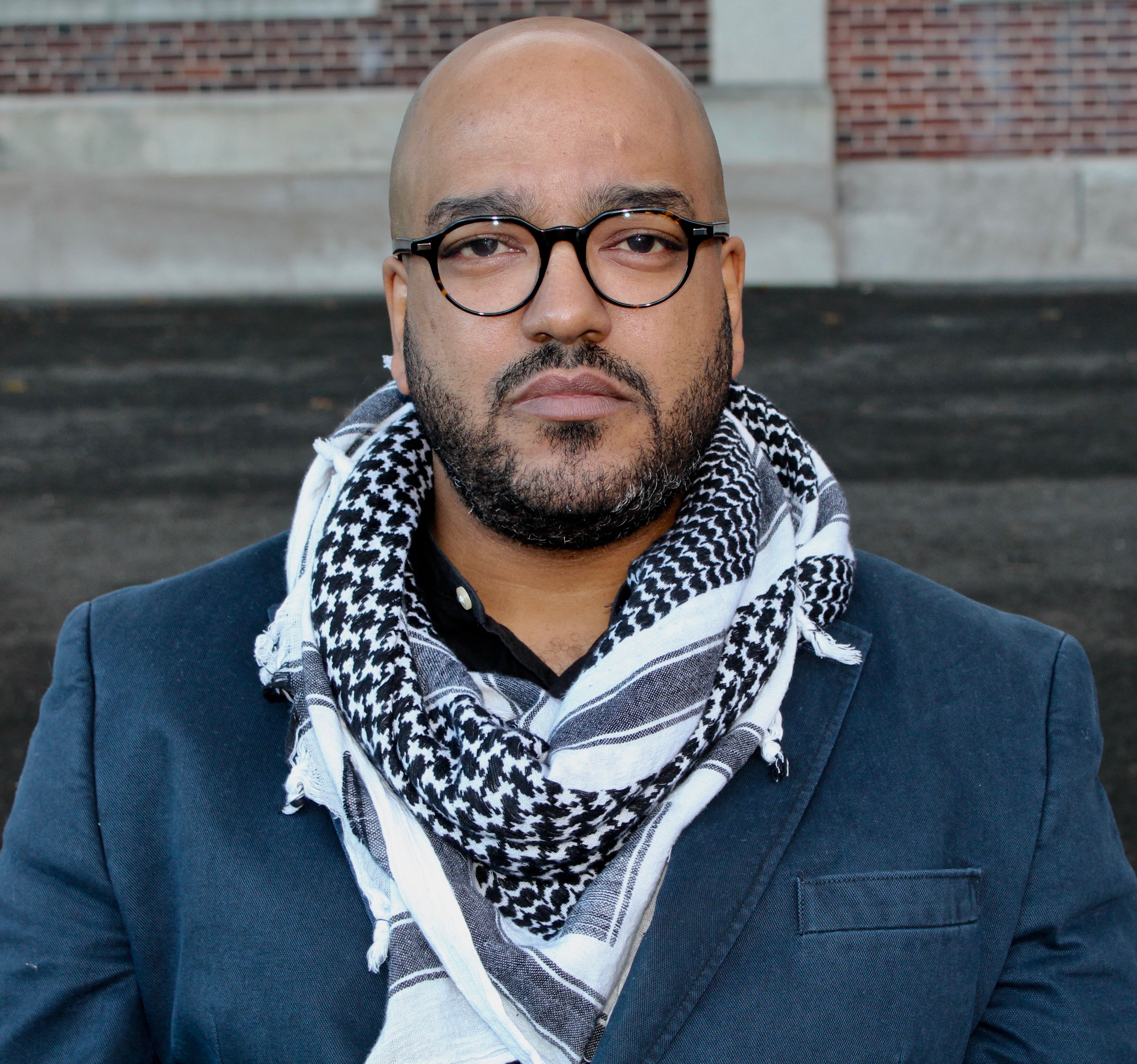 Photo of author staring seriously into the camera, wearing glasses, a blazer, and a keffiyeh. There is a brick wall in the background.