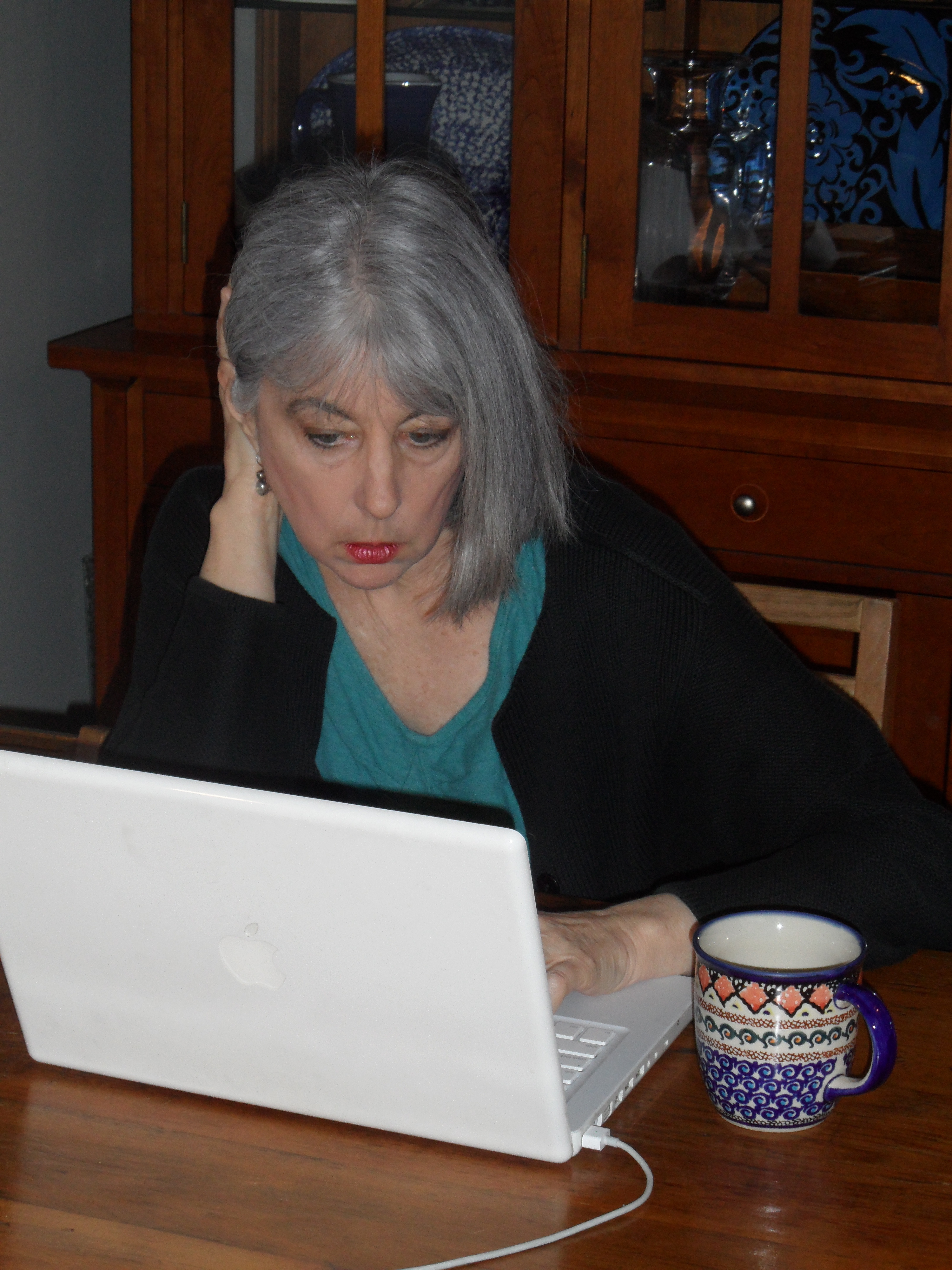 Jan Carr sitting at home, sitting table working on computer, with a cup of coffee at the ready.