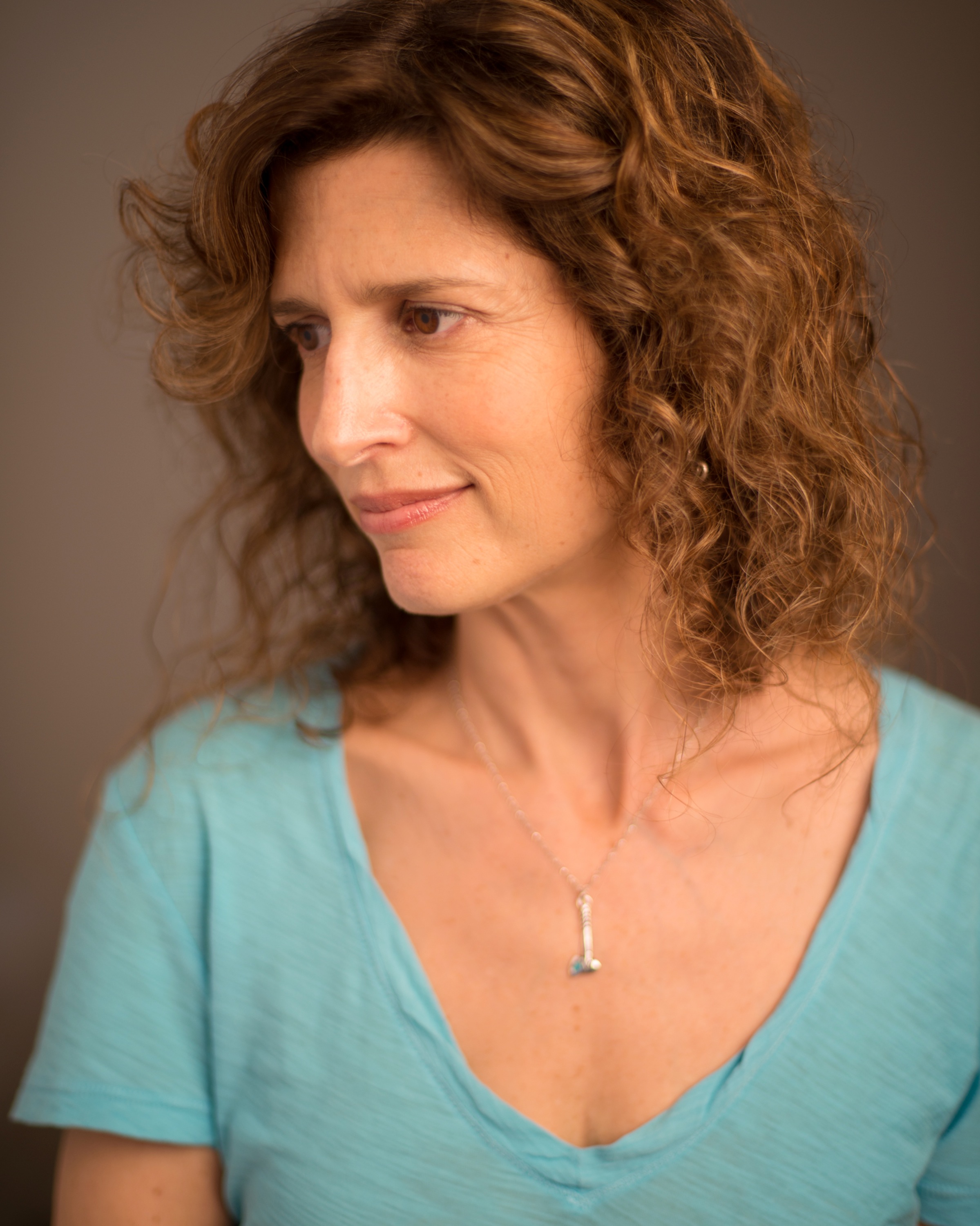A headshot of a woman with brown wavy hair gazing to the left, wearing a light blue t-shirt. Not smiling.