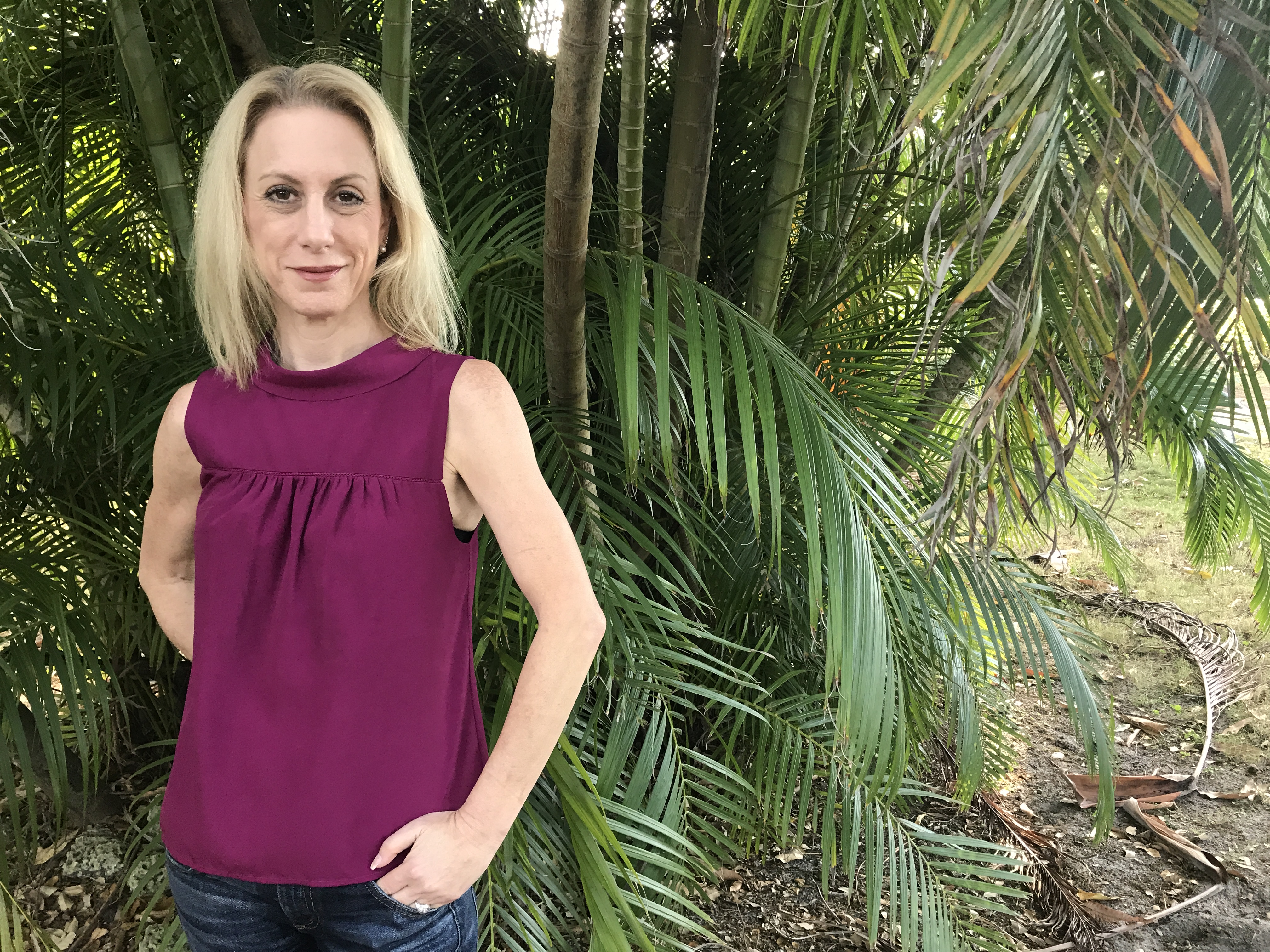 Jen is a Causasian woman with blond shoulder-length hair wearing a sleeveless magenta blouse. She is smiling and standing in front of tropical foliage.