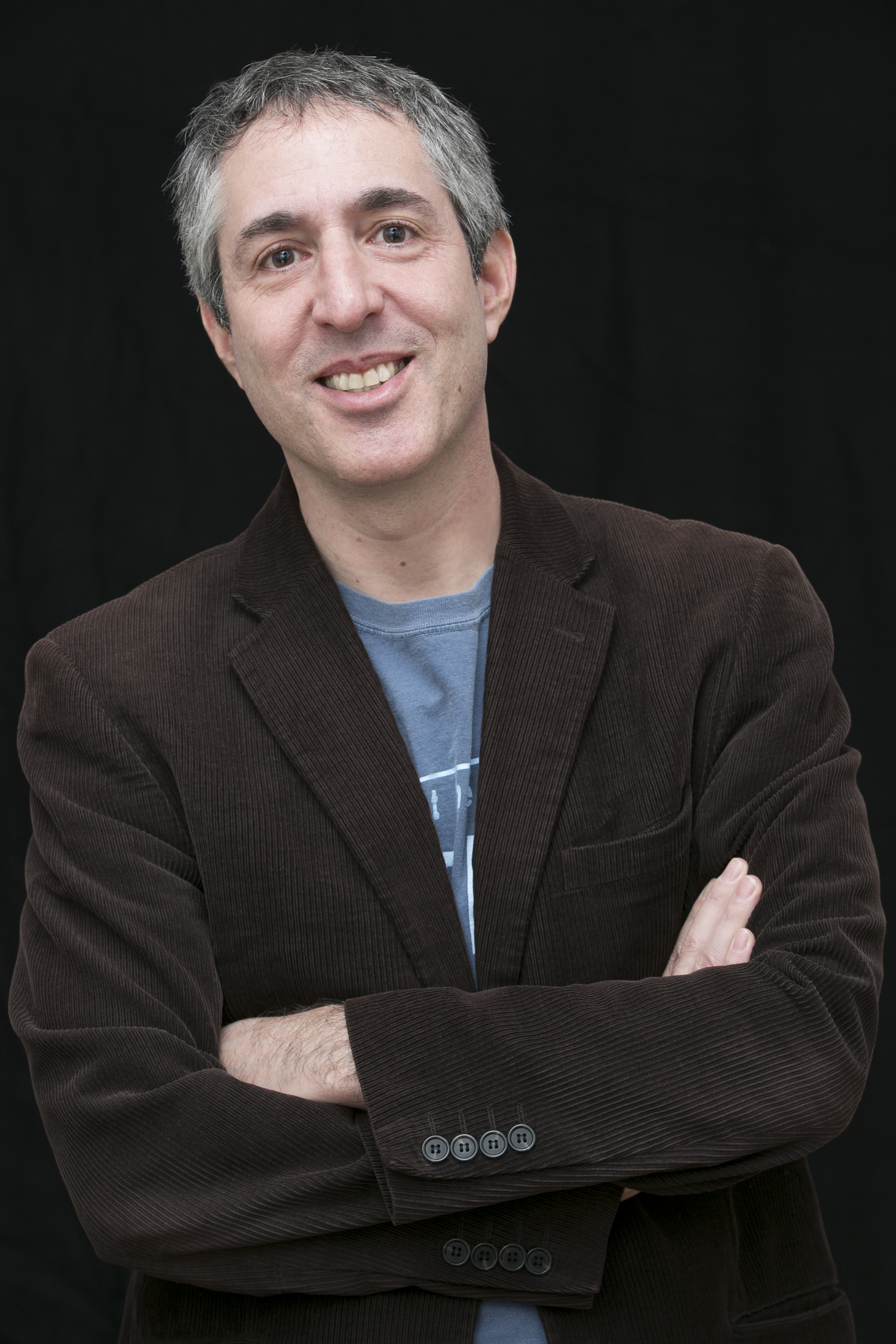 Photograph of Alex Zucker with his arms folded, in a brown corduroy jacket with a blue t-shirt on underneath