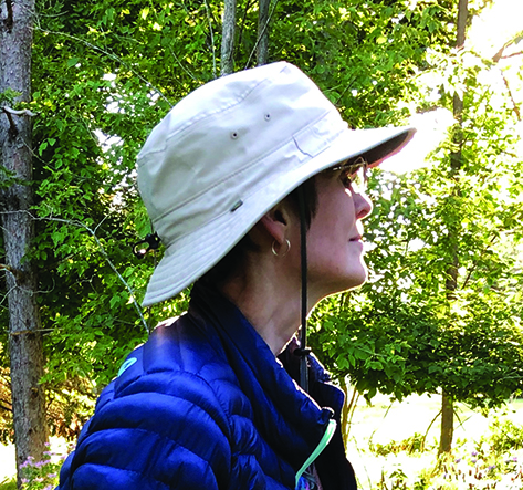 Woman with short brown hair wearing a hat looks off into the woods.