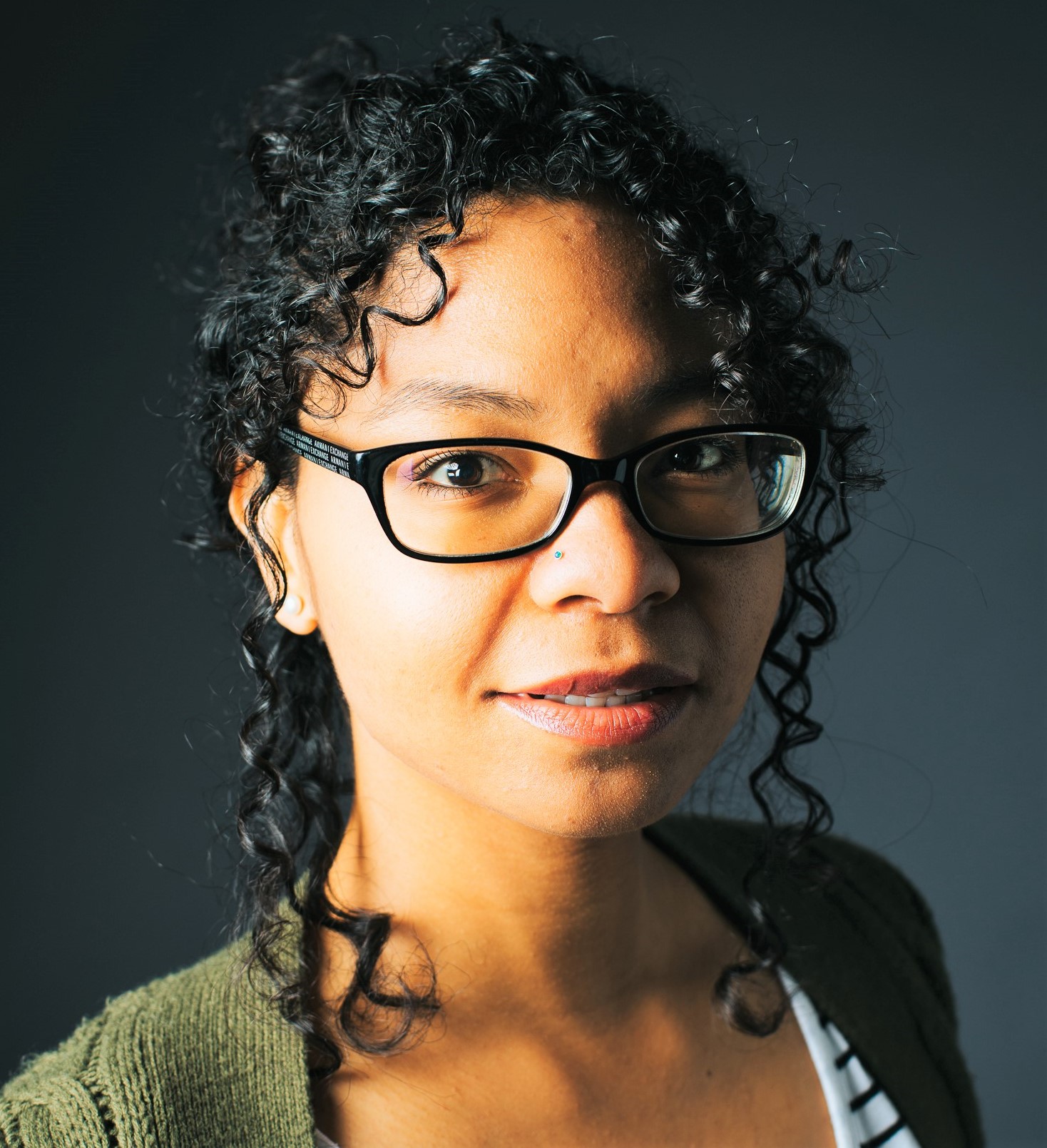 brown-skinned woman with curly brown hair and black glasses wearing a forest green cardigan and striped t-shirt