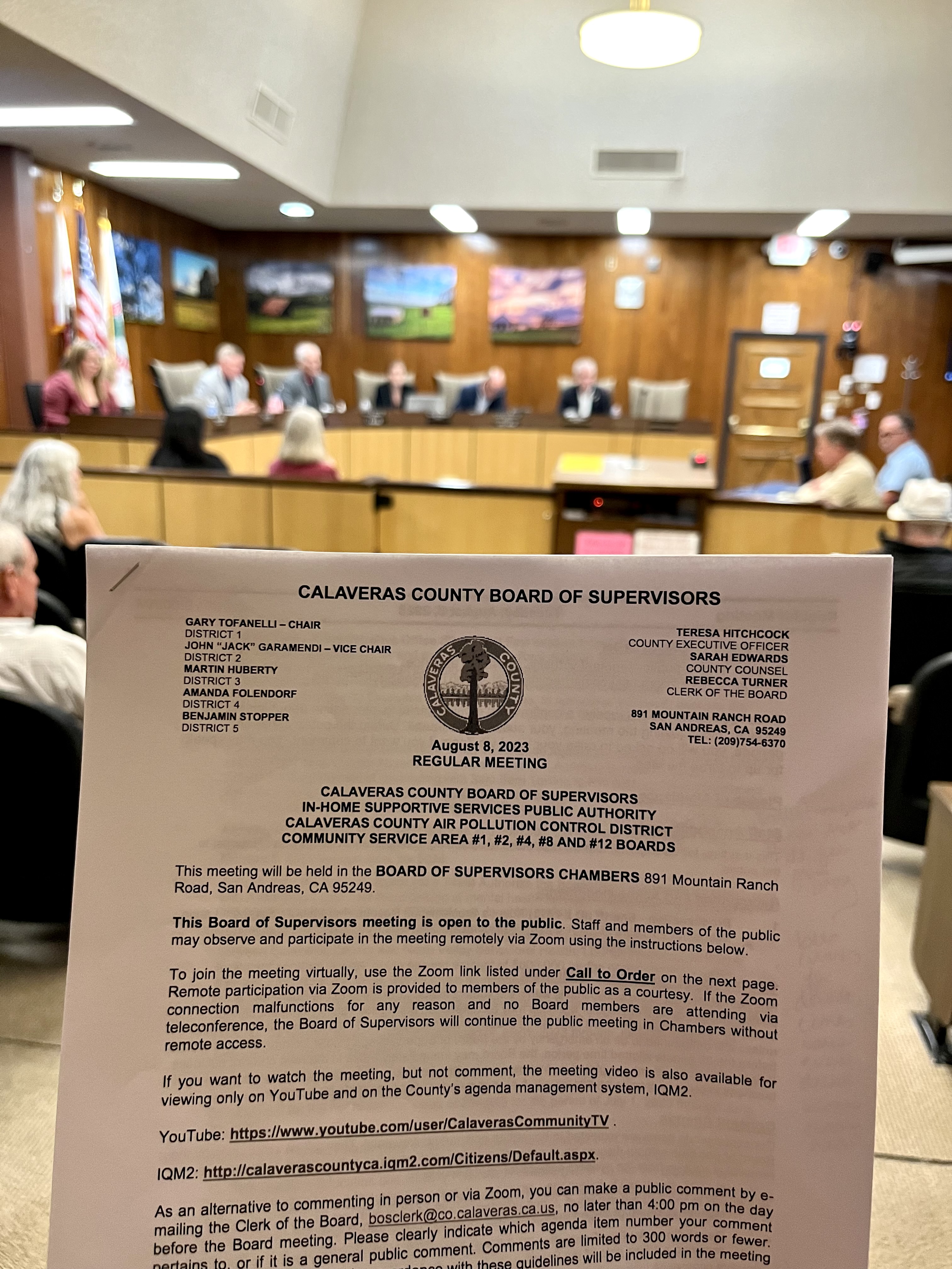 A paper copy of the Calaveras County Board of Supervisors agenda for the regular meeting is shown in focus inside the Board of Supervisors Chambers with the five member board blurred in the background.